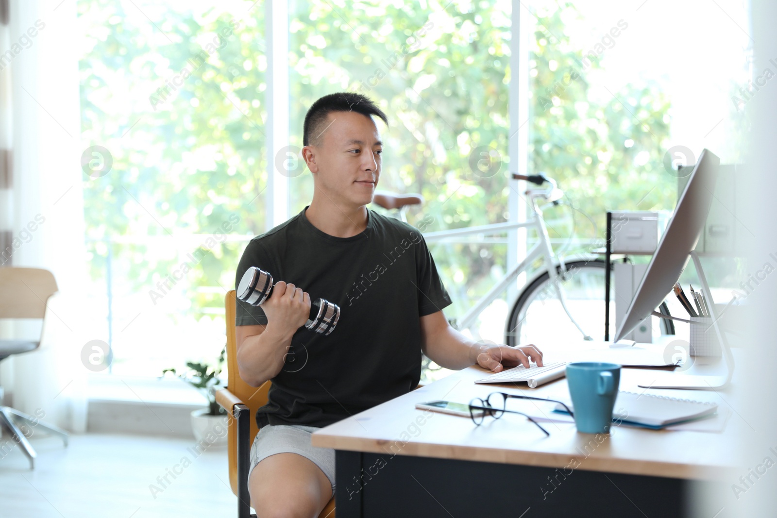 Photo of Young man lifting weights and using computer in office. Workplace fitness