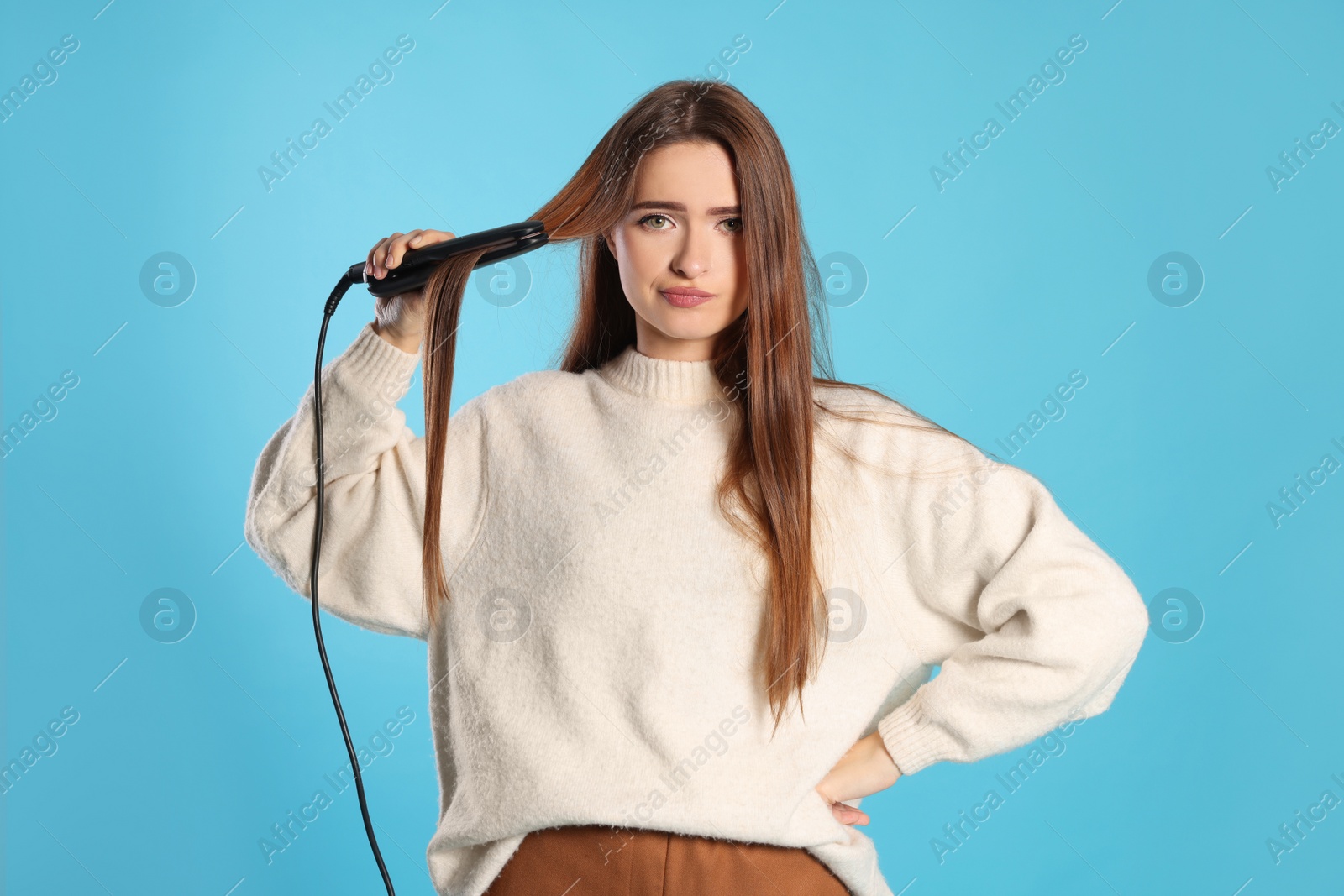 Photo of Upset young woman with flattening iron on light blue background. Hair damage