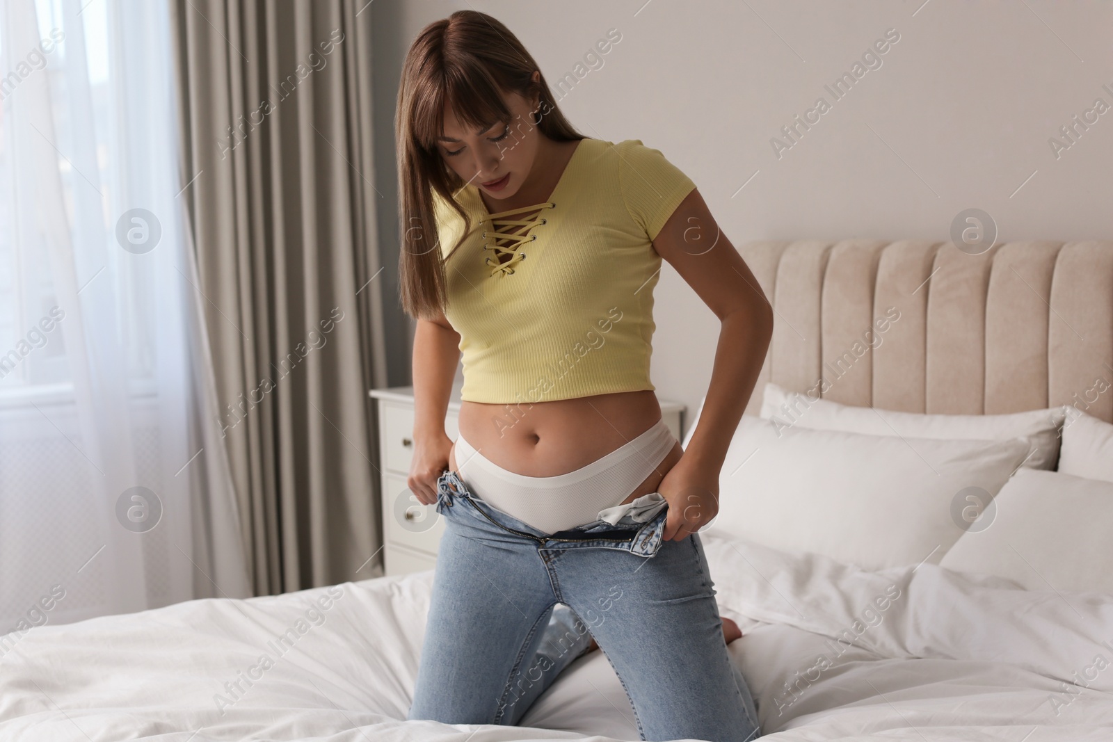 Photo of Young woman struggling to squeeze into tight jeans on bed at home