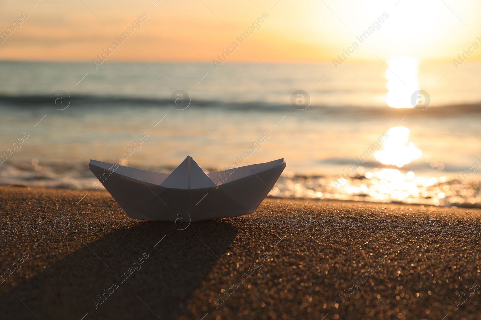 Photo of White paper boat on sand near sea at sunset, space for text