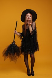 Photo of Young woman in scary witch costume with broom blowing kiss on orange background. Halloween celebration