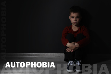 Sad little boy sitting alone near black wall, space for text. Autophobia - fear of isolation