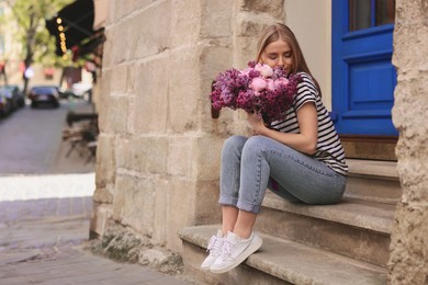 Beautiful woman with bouquet of spring flowers on stairs near building, space for text