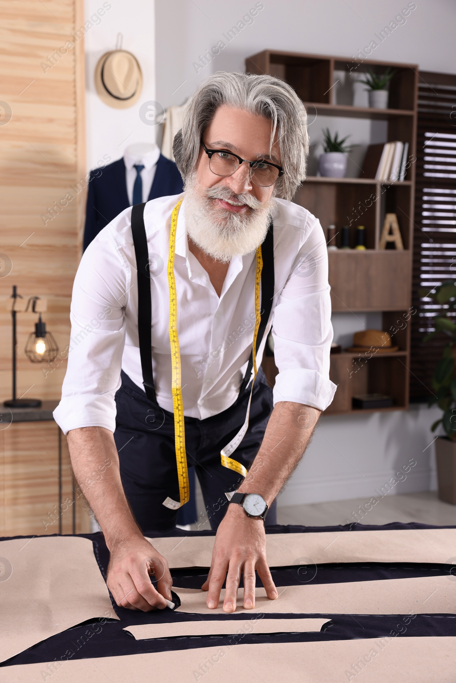 Photo of Professional tailor marking sewing pattern on fabric with chalk at table in workshop