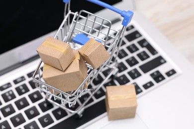 Photo of Internet store. Small cardboard boxes, shopping cart and laptop on table, closeup