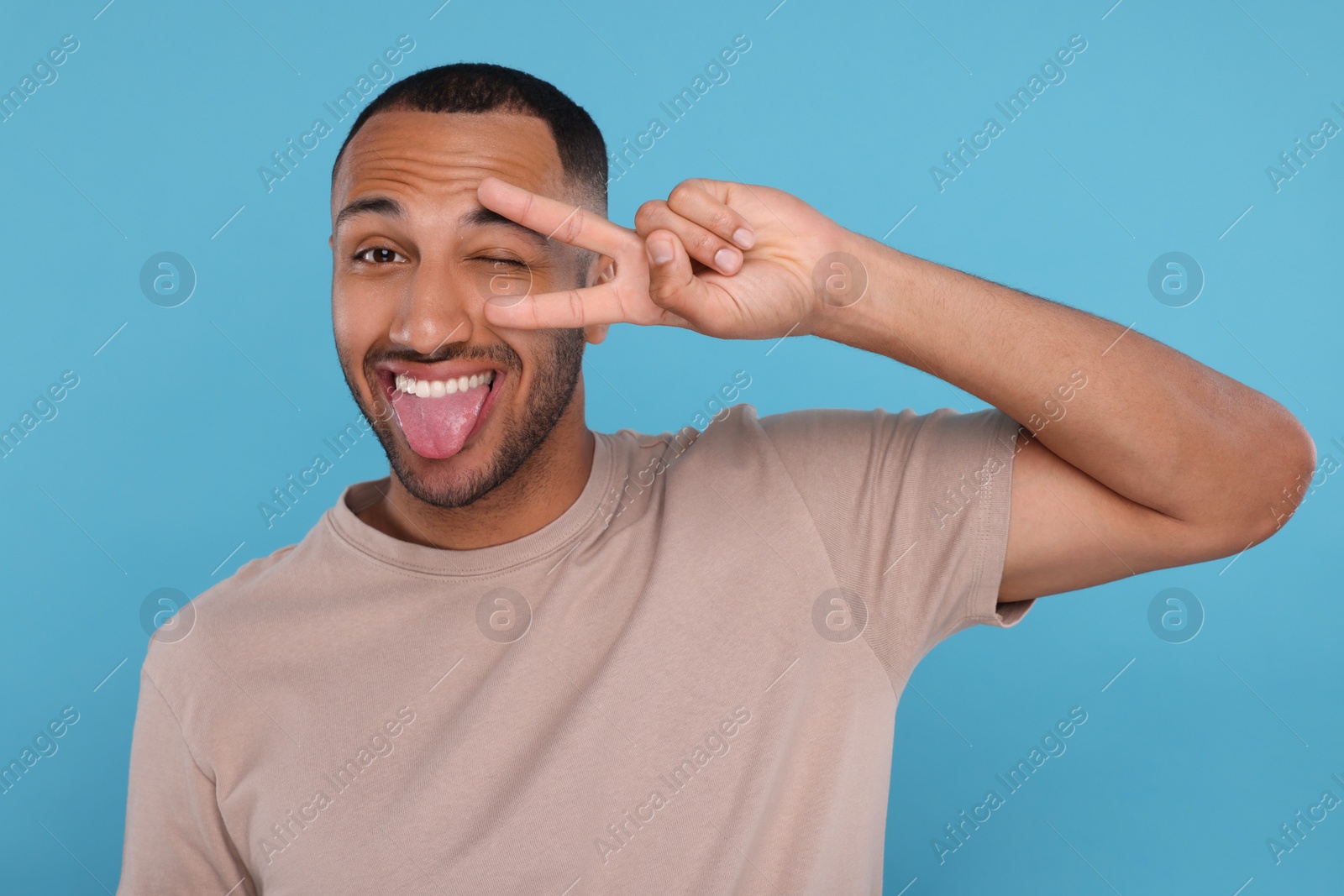 Photo of Happy young man showing his tongue and V-sign on light blue background