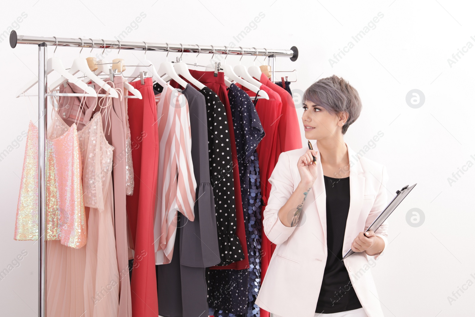 Photo of Beautiful young stylist holding clipboard near rack with designer clothes on white background