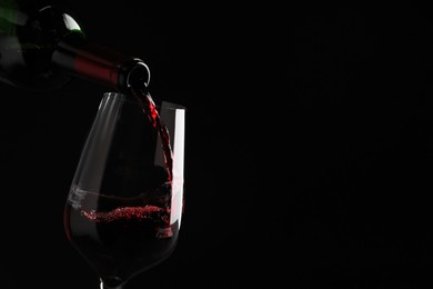 Pouring tasty red wine from bottle into glass on black background, closeup. Space for text