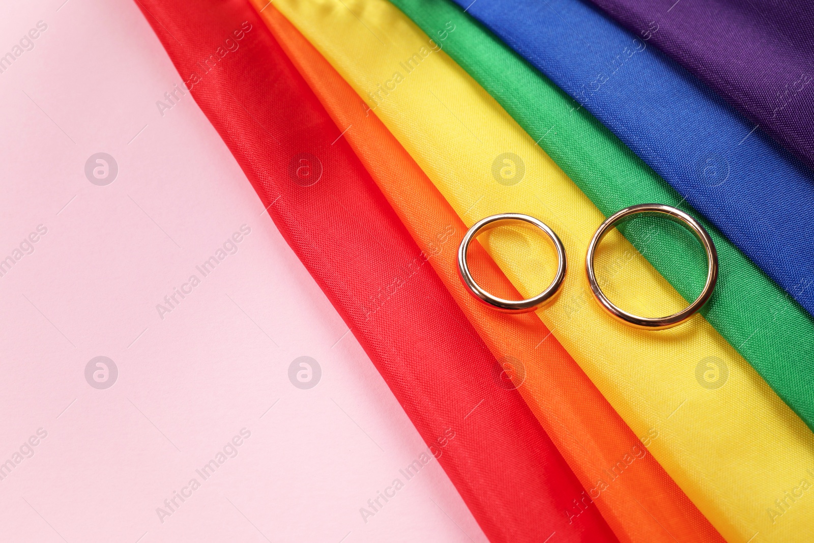 Photo of Rainbow LGBT flag and wedding rings on pink background, top view. Space for text