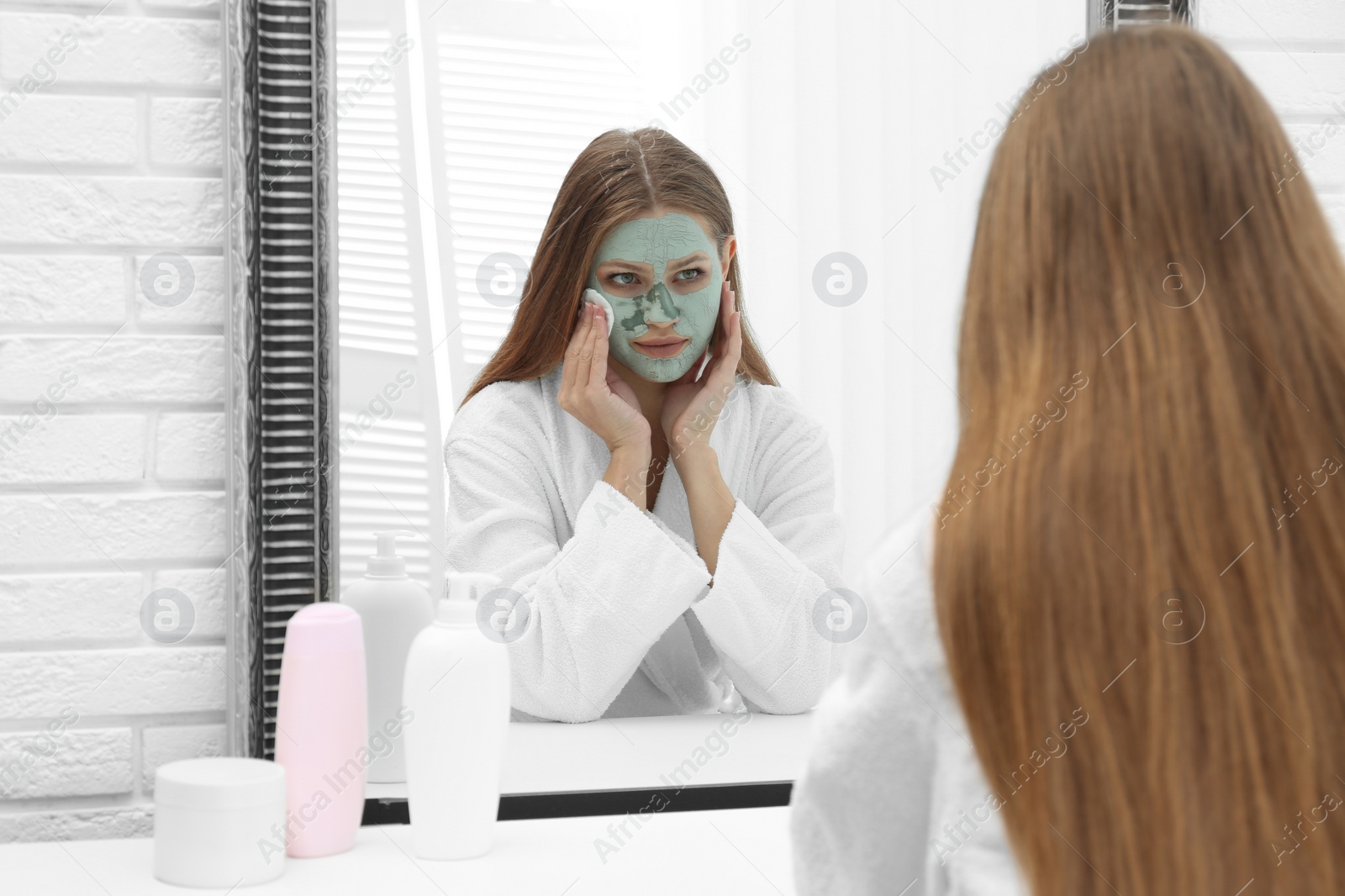 Photo of Beautiful woman removing homemade clay mask from her face at mirror indoors