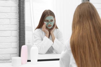 Beautiful woman removing homemade clay mask from her face at mirror indoors