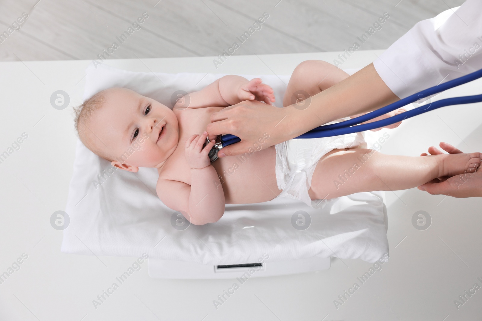 Photo of Pediatrician weighting and examining cute little baby with stethoscope in clinic, closeup