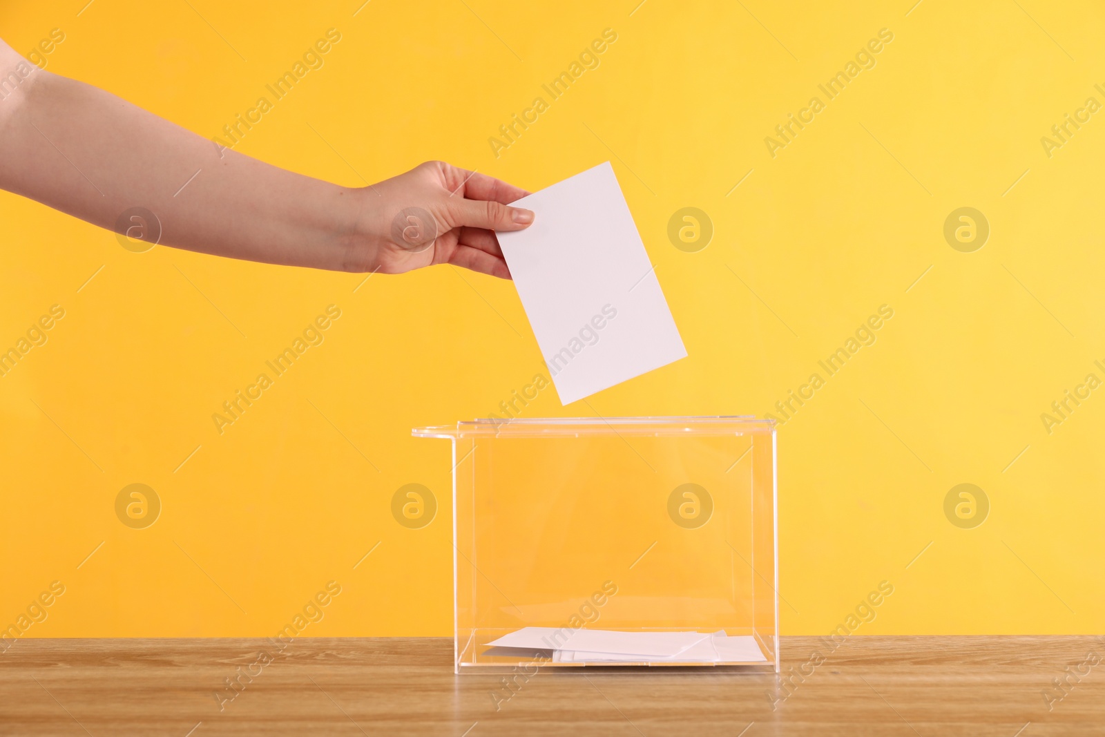 Photo of Woman putting her vote into ballot box on wooden table against orange background, closeup