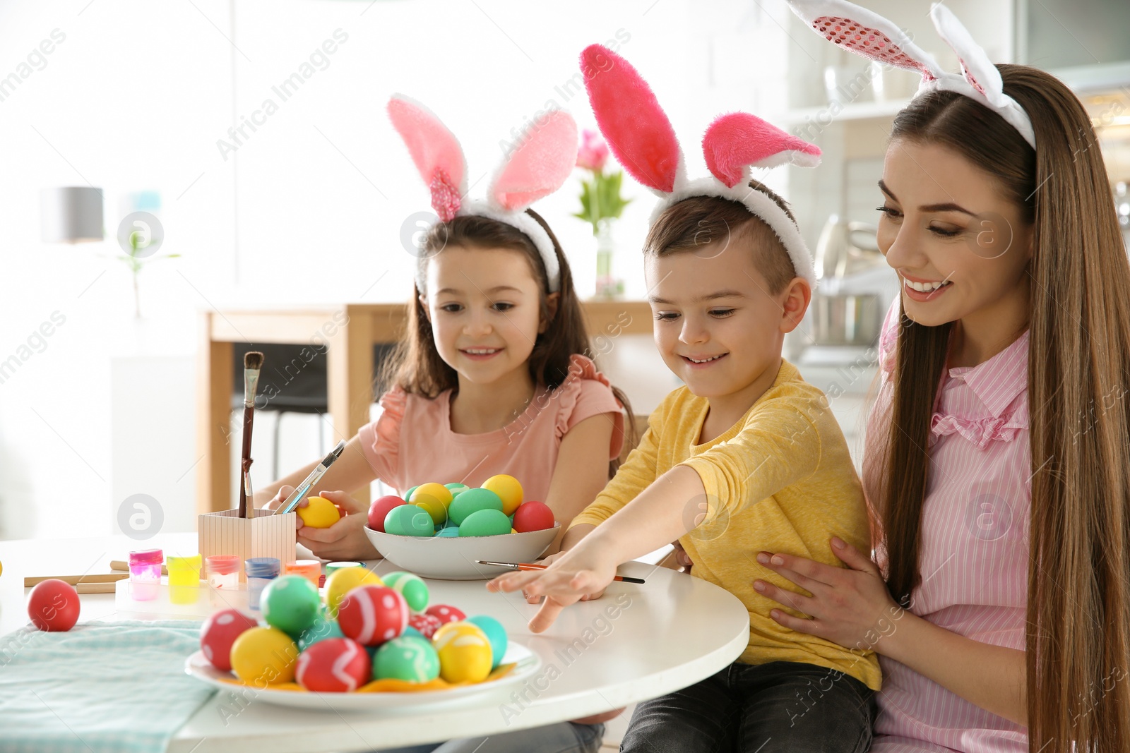 Photo of Mother and her children with bunny ears headbands painting Easter eggs in kitchen, space for text