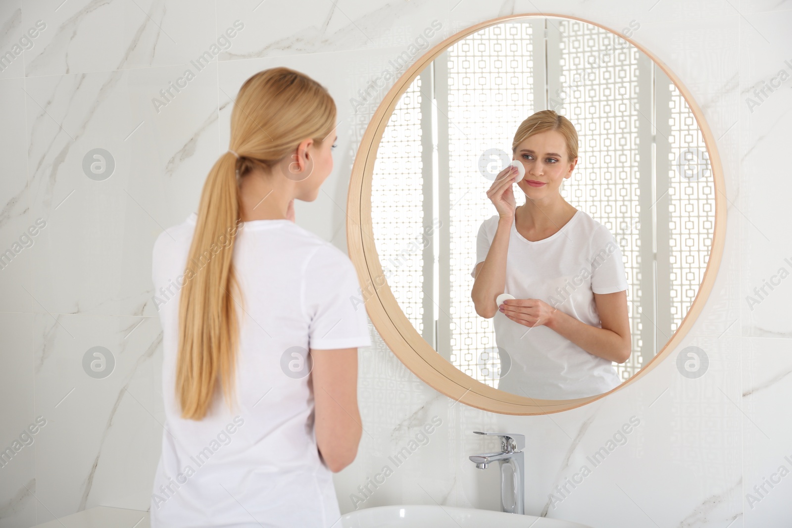 Photo of Happy young woman cleaning face with cotton pad near mirror in bathroom