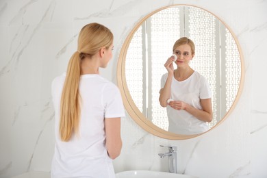 Happy young woman cleaning face with cotton pad near mirror in bathroom