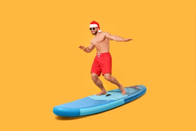 Happy man in Santa hat balancing on SUP board against orange background