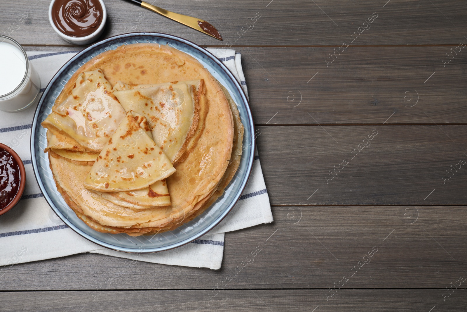 Photo of Delicious crepes served with chocolate paste, jam and milk on wooden table, flat lay. Space for text