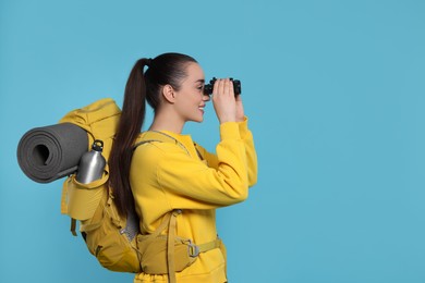 Photo of Young woman with backpack looking through binoculars on light blue background, space for text. Active tourism