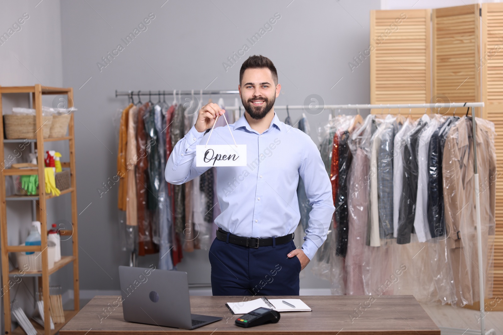Photo of Dry-cleaning service. Happy worker holding Open sign at workplace indoors
