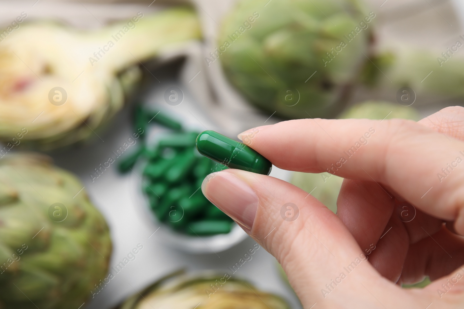 Photo of Woman holding pill over table with fresh artichokes, closeup