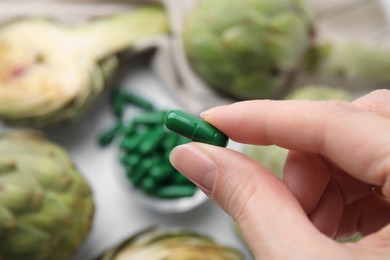 Woman holding pill over table with fresh artichokes, closeup