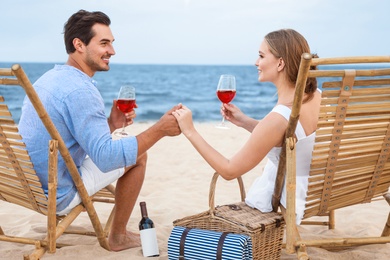 Happy young couple with glasses of wine sitting on deck chairs at sea beach