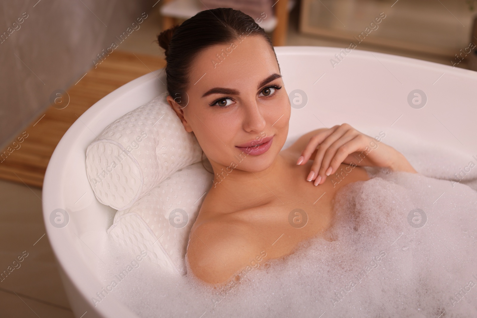 Photo of Young woman using pillow while enjoying bubble bath indoors