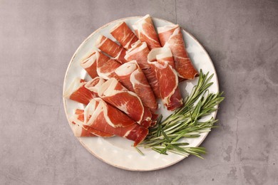 Photo of Rolled slices of delicious jamon with rosemary on grey table, top view