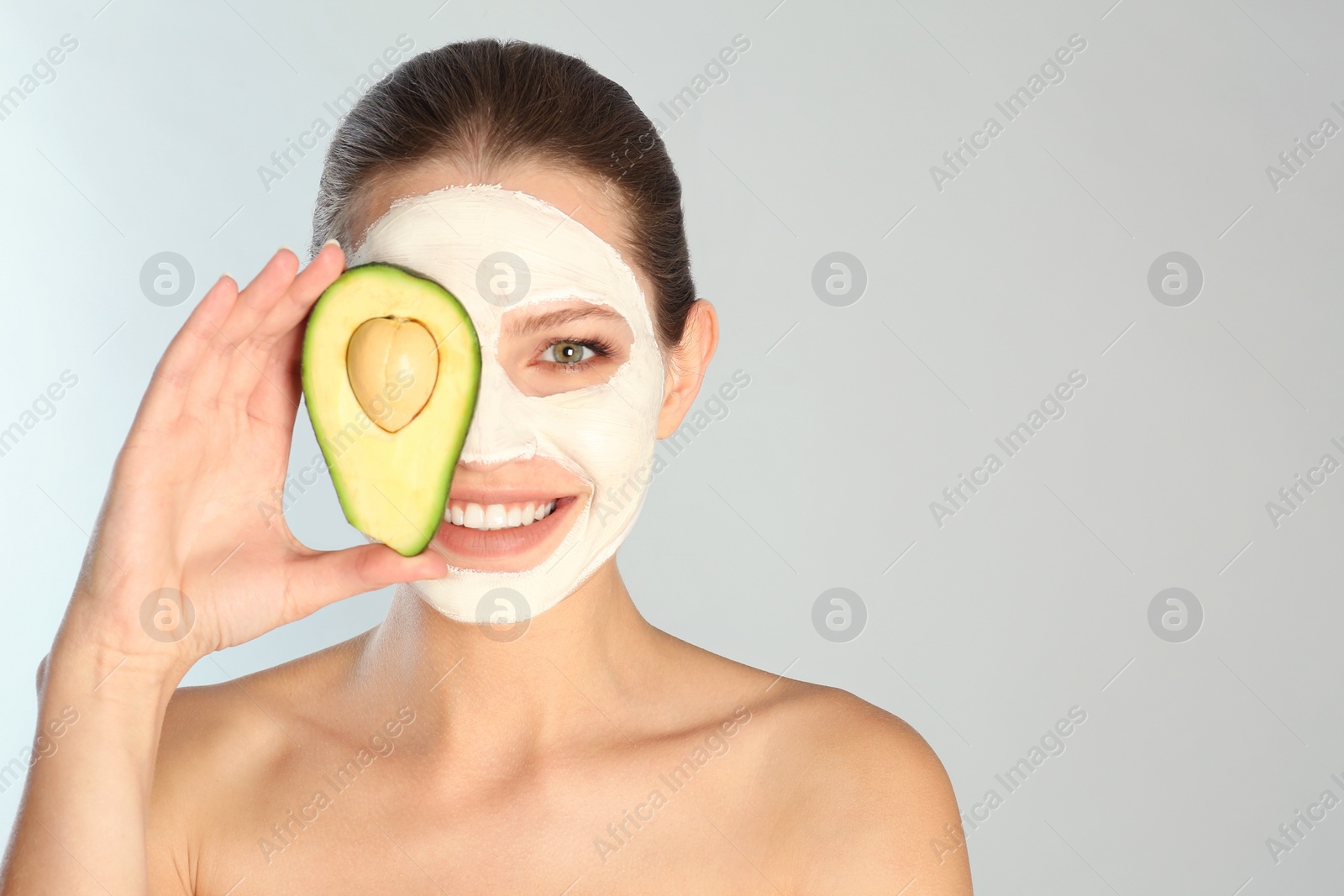 Photo of Beautiful woman holding avocado near her face with clay mask against grey background. Space for text