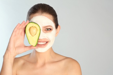 Photo of Beautiful woman holding avocado near her face with clay mask against grey background. Space for text