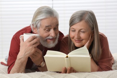 Photo of Senior man with cup of drink and his wife reading book on bed at home