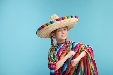 Cute girl in Mexican sombrero hat and poncho dancing on light blue background