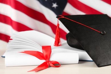 Photo of Black graduation hat, diploma and book on white wooden table against American flag