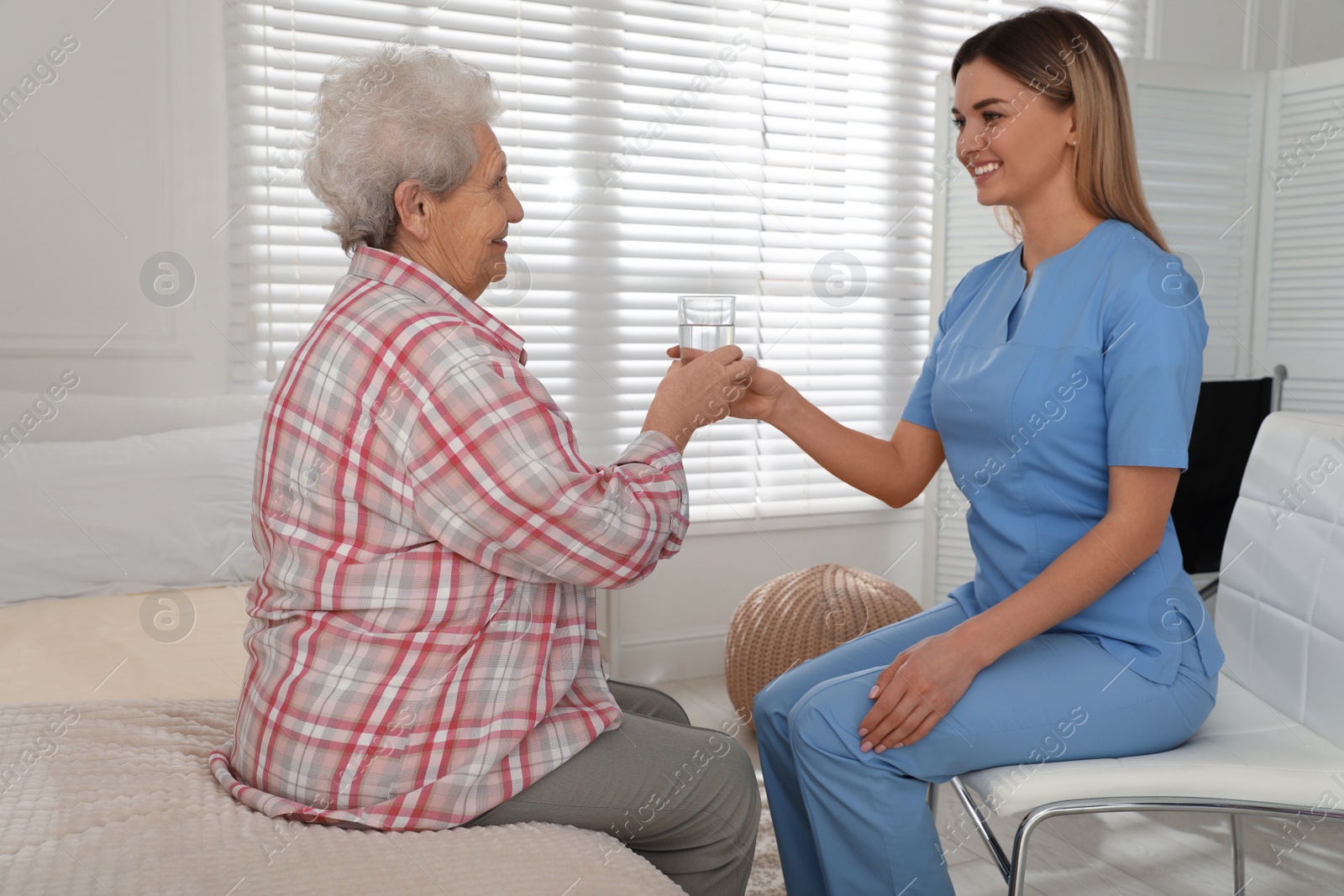 Photo of Young caregiver giving water to senior woman in room. Home care service