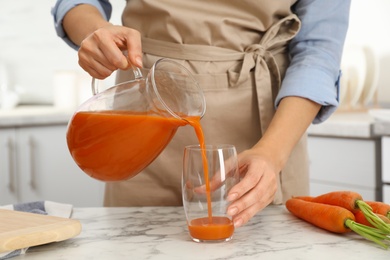 Woman pouring tasty carrot juice from jug into glass at table indoors, closeup