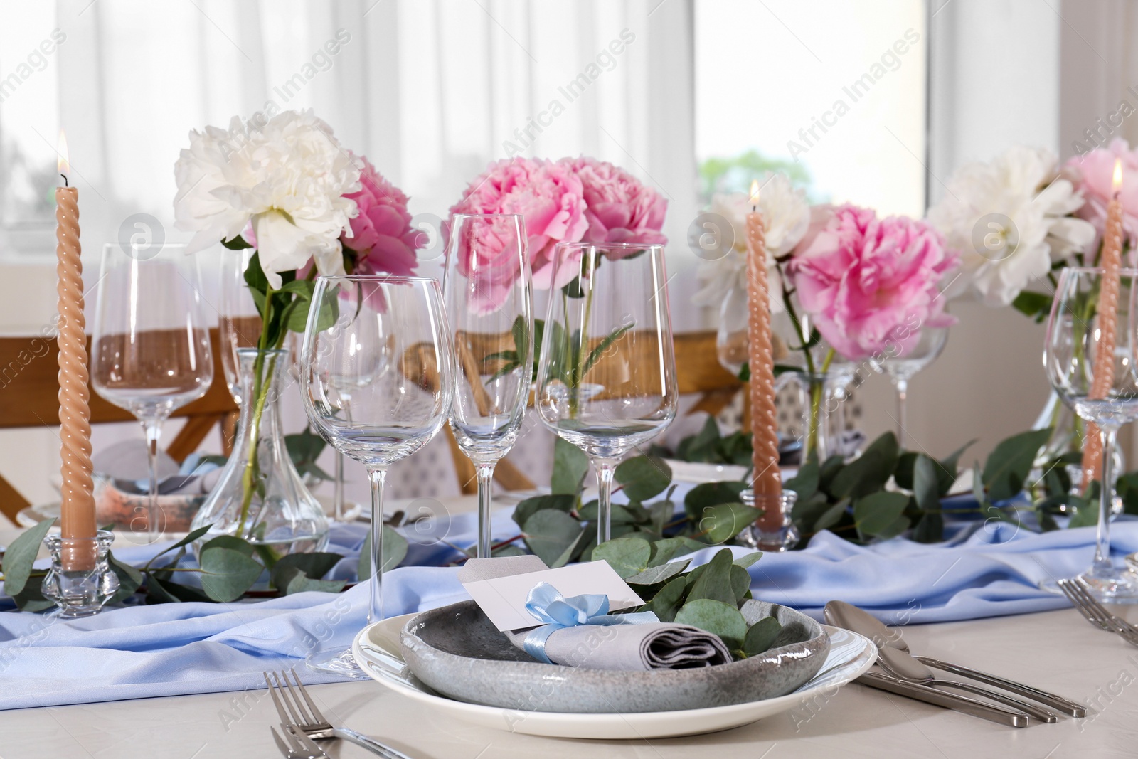 Photo of Beautiful table setting. Plate with greeting card, napkin and branch near glasses, peonies, burning candles and cutlery on table in room