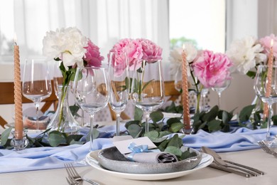 Photo of Beautiful table setting. Plate with greeting card, napkin and branch near glasses, peonies, burning candles and cutlery on table in room