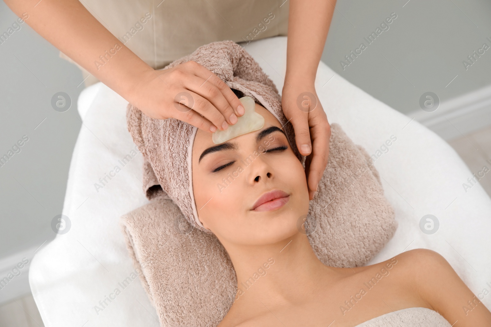 Photo of Young woman receiving facial massage with gua sha tool in beauty salon