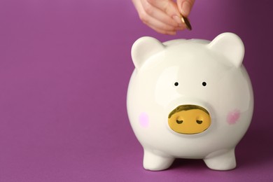 Photo of Woman putting coin into piggy bank on purple background, closeup. Space for text