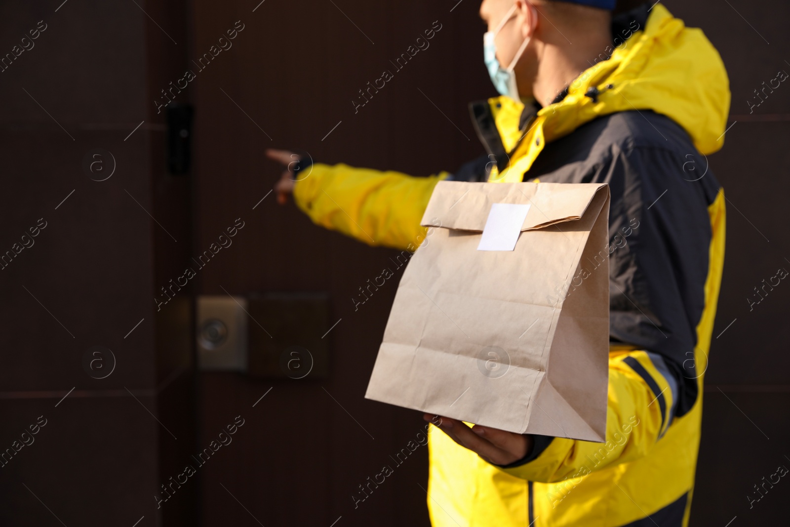 Photo of Courier in medical mask ringing doorbell outdoors, focus on paper bag with takeaway food. Delivery service during quarantine due to Covid-19 outbreak