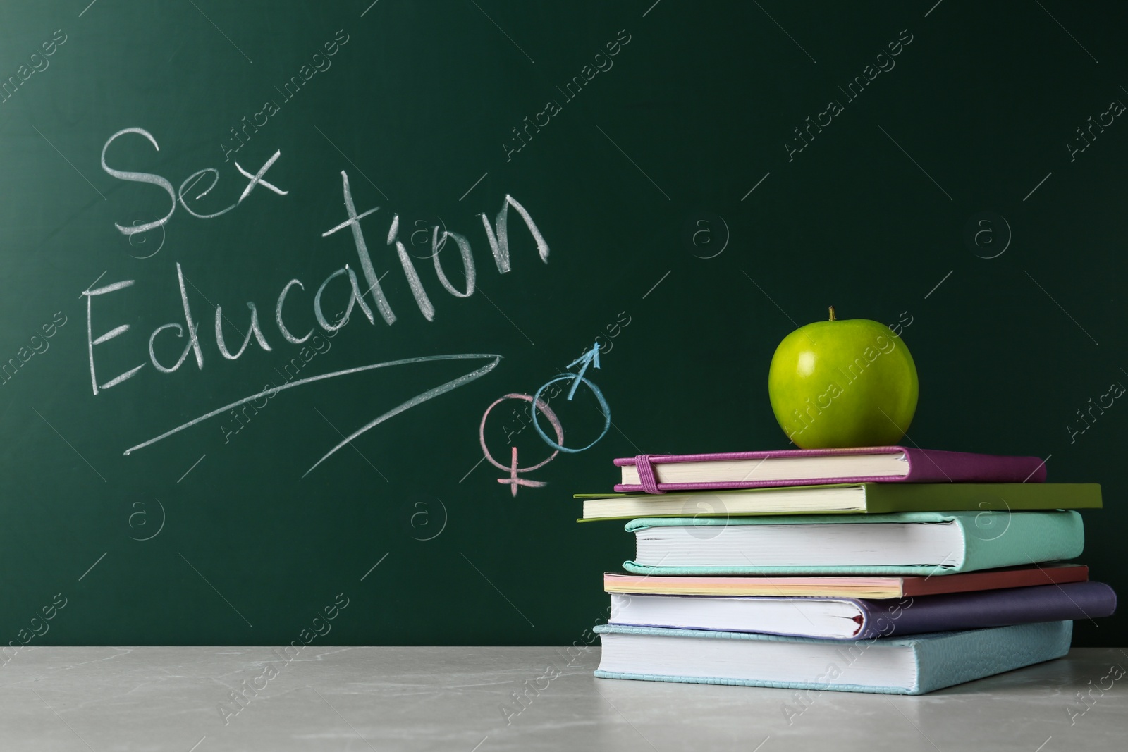 Photo of Books and apple on grey table near chalkboard with phrase "Sex education"