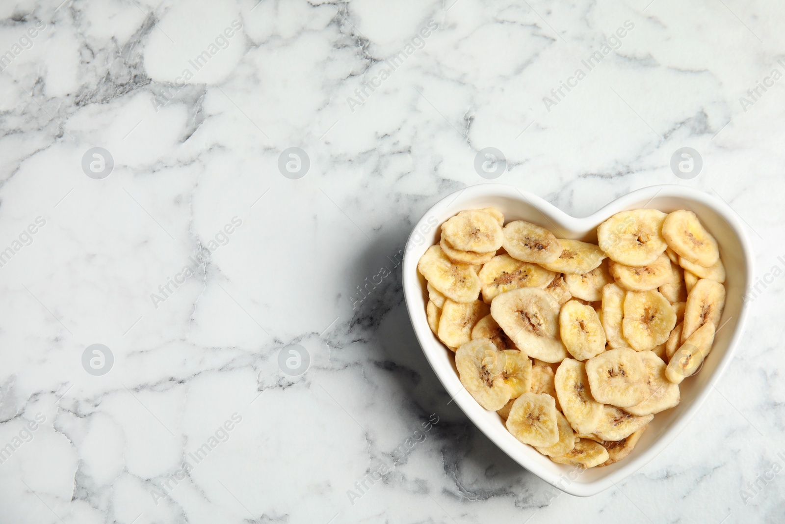 Photo of Heart shaped plate with banana slices on marble table, top view with space for text. Dried fruit as healthy snack