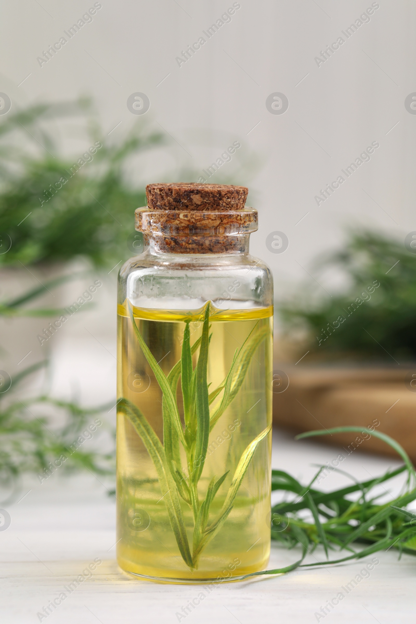 Photo of Bottle of essential oil and fresh tarragon leaves on white wooden table