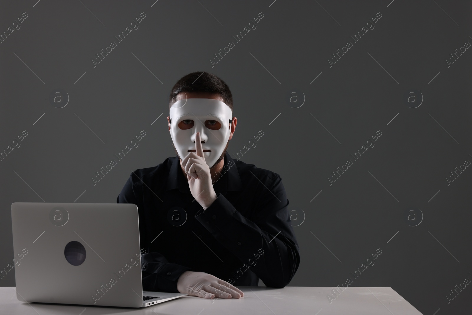 Photo of Man in mask and gloves sitting with laptop and showing hush gesture at white table against grey background. Space for text