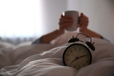 Woman with cup in bed, focus on alarm clock. Morning time