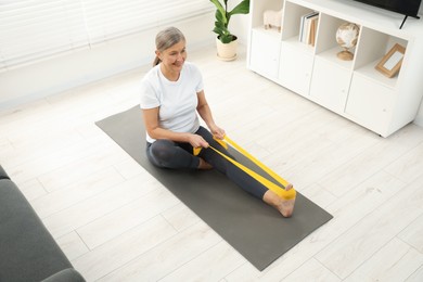 Photo of Senior woman doing exercise with fitness elastic band on mat at home