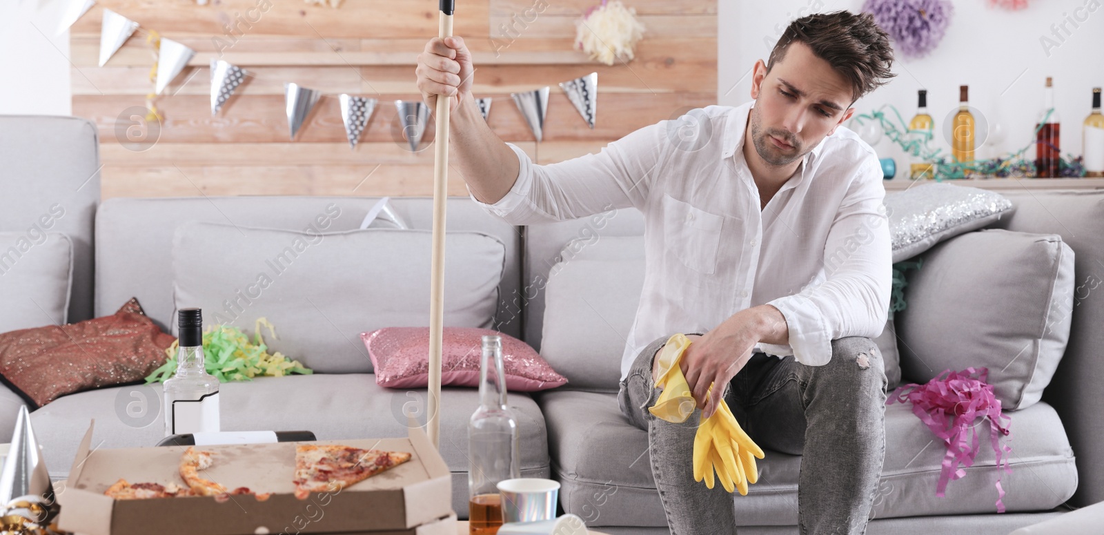 Image of Young man with mop suffering from hangover in messy room after party. Banner design