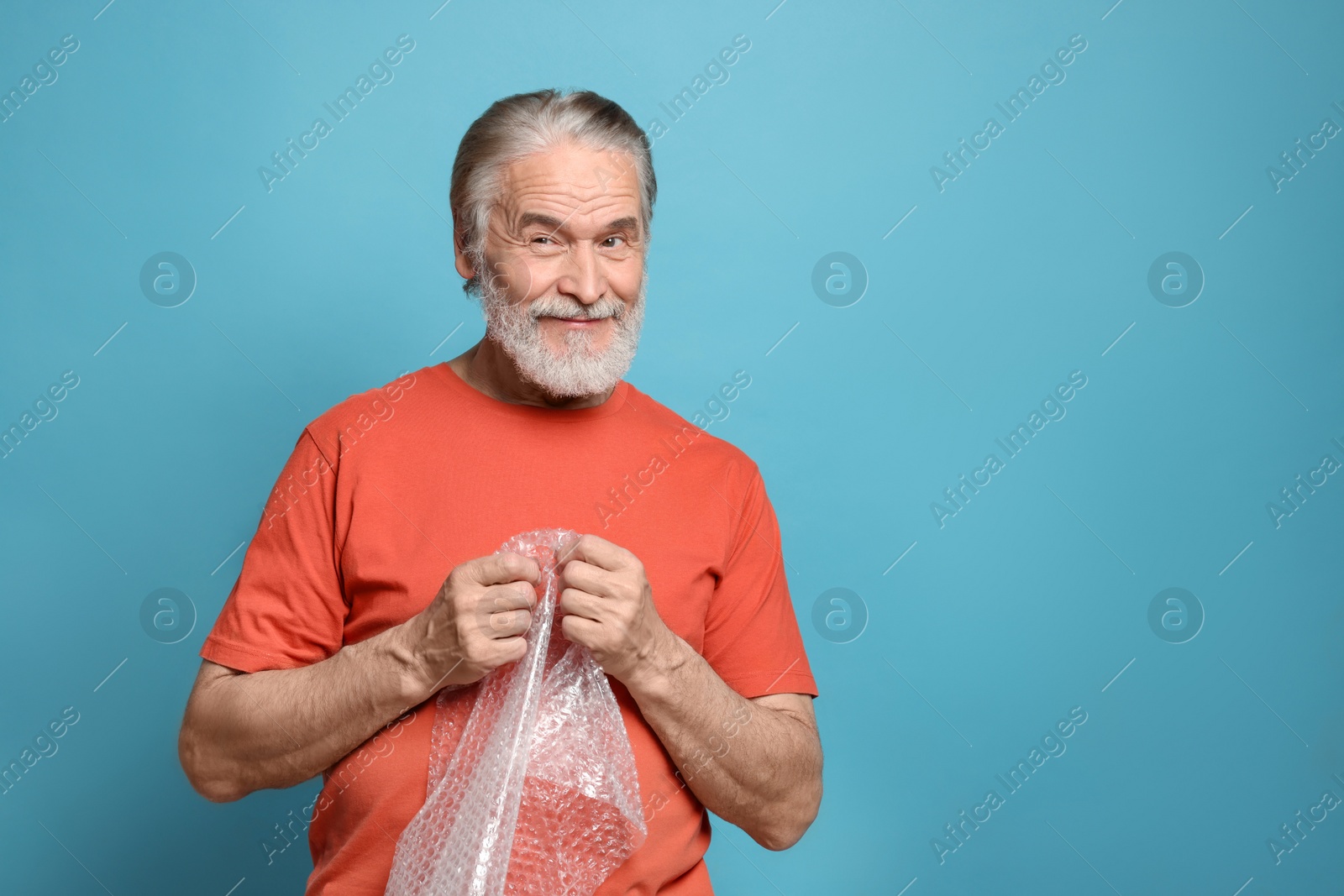 Photo of Happy senior man popping bubble wrap on light blue background, space for text. Stress relief