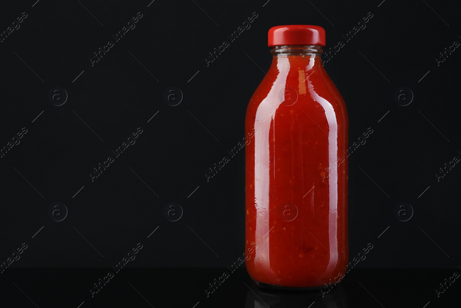 Photo of Spicy chili sauce in bottle against dark background, space for text